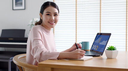 Wall Mural - Portrait headshot young attractive beautiful asia female sit on desk smile to camera work at home remotely computer laptop job. College student, employee feel cozy happy in weekend work life balance.