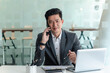 © amnaj - Asian businessman sitting on the phone with a customer with a laptop and document at his office desk.
