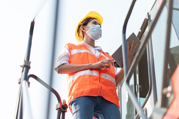 Female engineer wears a coronavirus mask on a crane to transport a container in the container yard. Working woman concept.