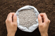 Young adult woman hands holding opened plastic bag with gray complex fertiliser granules on dark soil background. Closeup. Product for root feeding of vegetables, flowers and plants. Top down view.