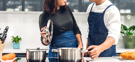 Wall Mural - Young asian family couple having fun standing near stove and cooking together.Happy couple looking and smelling tasting fresh delicious from soup in a pot with steam at white interior kitchen