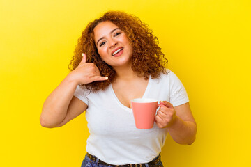 Wall Mural - Young latin curvy woman holding a cup isolated on yellow background showing a mobile phone call gesture with fingers.