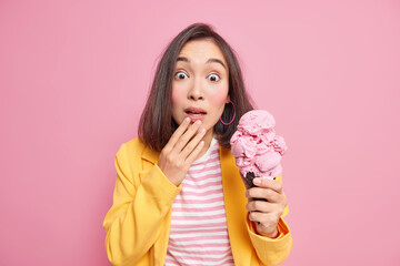 Wall Mural - Photo of surprised young Asian woman with dark hair stares shocked dressed in striped t shirt and yellow jacket holds yummy ice cream isolated over pink background. People and emotions concept