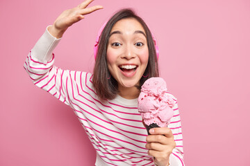 Wall Mural - Indoor shot of brunette woman with eastern appearance dances carefree raises arm smiles gladfully eats delicious ice cream listens music wears casual striped jumper isolated over pink background.