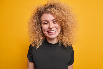 Portrait of good looking young European woman with curly bushy hair dressed in casual black t shirt smiles broadly expresses positive emotions isolated over yellow background laughs with joy
