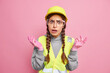 Indignant displeased woman builder industrial worker shrugs shoulders feels puzzled spreads hands sideways wears protective hardhat glasses reflective vest works at factory or construction site