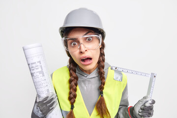 Construction industry. Puzzled surprised woman engineer with pigtails looks shocked at camera has engineering career has busy hard working day holds blueprint and tape measure comes on building site