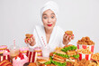 Surprised cheerful Asian woman focused at camera consumes too much fast food suffers calorie overload has giant portions eats fatty sandwiches burgers and cakes drinks milk cocktails poses indoor