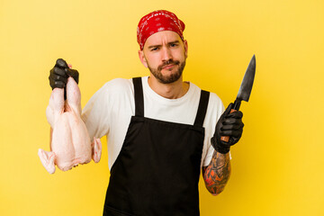 Wall Mural - Young tattooed batcher caucasian man holding a chicken isolated on yellow background