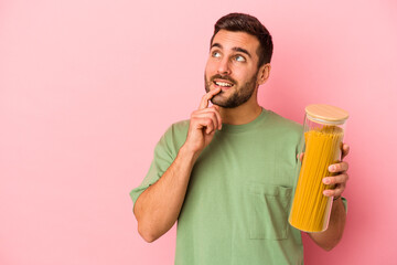 Wall Mural - Young caucasian man holding pasta jar isolated on pink background relaxed thinking about something looking at a copy space.