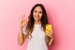 © Asier - Young mexican woman holding a mobile phone isolated on pink background cheerful and confident showing ok gesture.