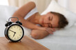 © volha_r - Beautiful young woman sleeping and smiling while lying in bed comfortably and blissfully on the background of alarm clock is going to ring. Sunbeam dawn on her face.