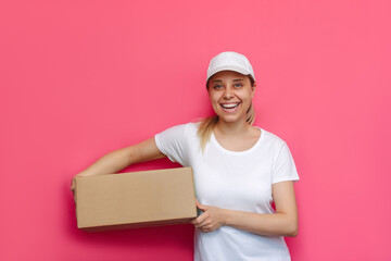 A young caucasian beautiful happy excited smiling blonde woman courier in a white t-shirt and cap holds a cardboard box isolated on a bright color pink background. A delivery girl delivers the package