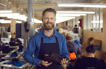 Portrait of footwear factory worker with brand new product. Happy young man standing in workshop, holding new leather boots, looking at camera and smiling. Shoe manufacturing industry concept