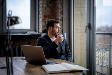 Handsome young brunette man businessman in a jacket sits in an office at a table with a laptop and papers