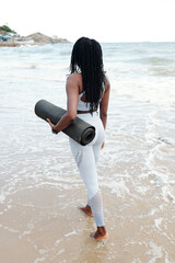 Wall Mural - Fit young sportswoman walking on beach with yoga mat in hands, enjoying beautiful view and breeze, view from the back