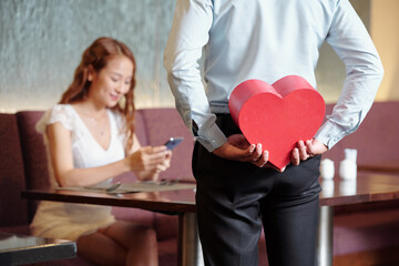 Wall Mural - Man hiding present in heart shape box for girlfriend who is sitting at cafe table and checking text messages on smartphone