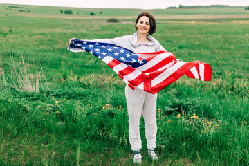 Extremely happy satisfied woman with red lips holding in hands big american flag sincerely rejoicing. Green field on the background. Patriotism