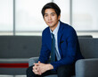 © Bangkok Click Studio - 20s visionary young handsome Asian executive businessman wearing a formal suit, sitting cross-legged on comfortable with relaxation, looking at camera with determination and confidence in success.