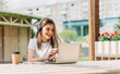 © Anna - Young hipster Woman in headphones using laptop, having a video call chat outside in park. Happy and smiling typing at notebook with coffee cup. Distance learning online education and work.