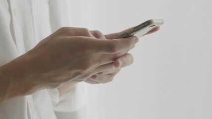 Close up hands young man using smartphone typing message chatting with friends on social media while standing beside the window at home.