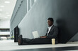Graphic full length portrait of African-American businessman using laptop while sitting on floor in office hall, copy space