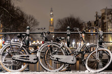 Winter Cityscape With Night View Of Snowy Covered Bicycles Parked On Canal Bridge, Blurred Architecture Traditional Canal Houses And The Westerkerk Church Tower, Prinsengracht, Amsterdam, Netherlands.