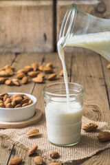 Wall Mural - Vegan almond milk pouring into a glass, almond kernels and whole almonds on the old rustic wooden table, vertical