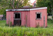 Old Pink And Salmon Painted Wooden Hut Type Storage Shed Used For Lawn Maintenance Tools And Farm Equipment With Open Windows And Shingle Roof Found In A Residential Backyard With Overgrown Grass.