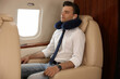 © New Africa - Young man with travel pillow resting while listening to music in airplane during flight