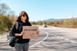 Smiling caucasian young woman in sunglasses and cap holding a cardboard sign with text anywhere. Copy space. The concept of local traveling and hitchhiking