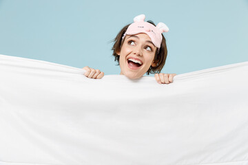 Wall Mural - Young dreamful happy woman in pajamas jam sleep eye mask rest relax at home hiding cover with white wrap under blanket look aside isolated on pastel blue background Good mood night bedtime concept