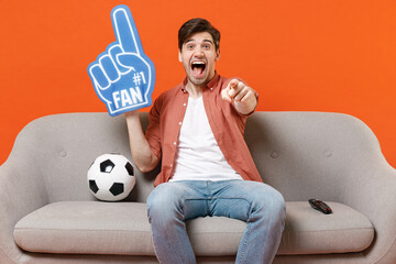 Wall Mural - Young man football fan in shirt foam glove finger support favorite team with soccer ball sit home sofa watch tv live stream point finger camera you isolated on orange background People sport concept.