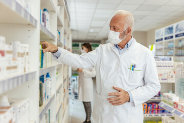 Wall Mural - Buying and selling medicines, doctor's advice and help to find medicines on the shelf. Close up of a man with gray hair in a white uniform and a protective face mask sorting drugs at the pharmacy