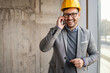 Smiling businessman with helmet on head standing next to a window in building in construction process and having business call.