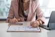 © amnaj - Close-up of a businesswoman holding a pencil pointing at a document with a smartphone at an office desk.
