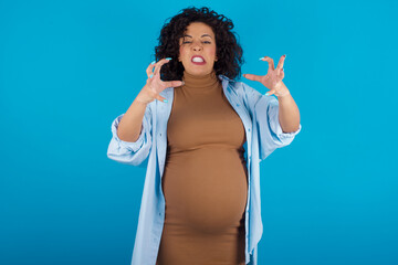 Canvas Print - young Arab pregnant woman wearing dress against blue wall Shouting frustrated with rage, hands trying to strangle, yelling mad.