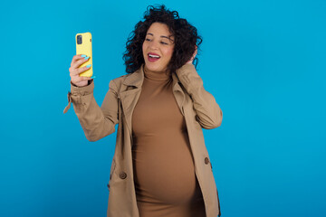 Sticker - young Arab pregnant woman wearing dress against blue wall smiling and taking a selfie ready to post it on her social media.