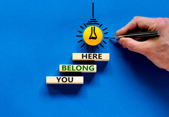 You belong here symbol. Wooden blocks with words 'You belong here' on beautiful blue background. Businessman hand, light bulb icon. Diversity, business, inclusion and belonging concept.