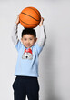portrait of preschool little smiling asian boy in trendy raglan with cool wolf print holding a basketball over his head. let's play
