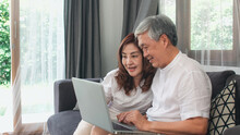 Asian Senior Couple Using Laptop At Home. Asian Senior Chinese Grandparents, Surf The Internet To Check Social Media While Lying On Sofa In Living Room At Home Concept.