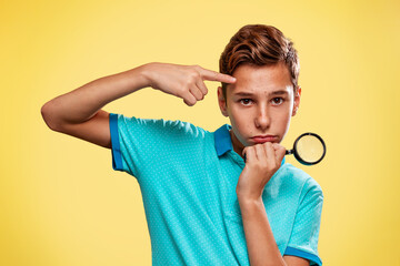 Wall Mural - A teenage boy in a blue t-shirt with pimples pointing on his face, holding a magnifying glass. Yellow background. Cosmetology, Dermatology and acne concept