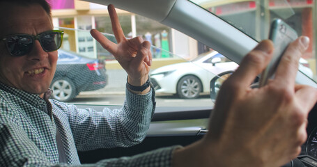 A man with glasses takes a selfie while driving a car. He holds the phone in his hands, takes photos. Starts moving on the car, gesture bye. Blogger's life.