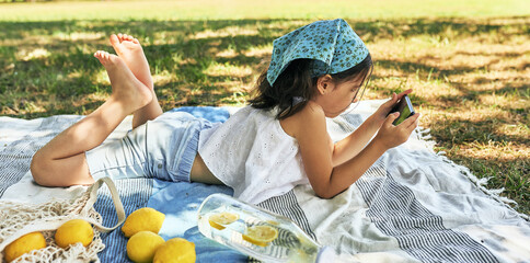 Wall Mural - Side view of a little girl lying on the blanket playing on the device in the park. Kid having cozy summer picnic with family with fresh lemonade and fruits outdoor.