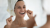 Fototapeta Do pokoju - Happy young woman wrapped in bath towel flossing white healthy teeth with thread after shower, looking at reflection in mirror, smiling with open mouth. Dental care, oral hygiene, routine concept
