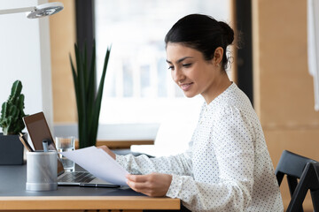 Smiling confident Indian businesswoman reading document, working on laptop, sitting at desk, young woman employee or student typing, holding paper, preparing financial report or research project