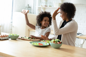 Wall Mural - Overjoyed young African American mom and small ethnic daughter give high five cook in kitchen together. Happy biracial mother and little girl child prepare healthy vegetarian diet food at home.