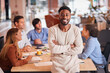 © Monkey Business - Portrait Of Businessman With Colleagues In Background Sitting Around Table In Open Plan Office