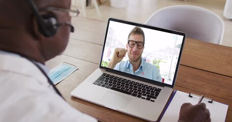 Wall Mural - African american male doctor wearing phone headset taking notes while having a video call on laptop