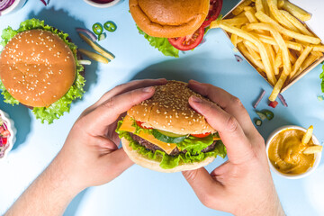 Wall Mural - Fastfood, burger party concept. Various delicious burgers set with french fries, Man hands hold tasty hamburgerm and sauces on bright blue background flatlay top view copy space